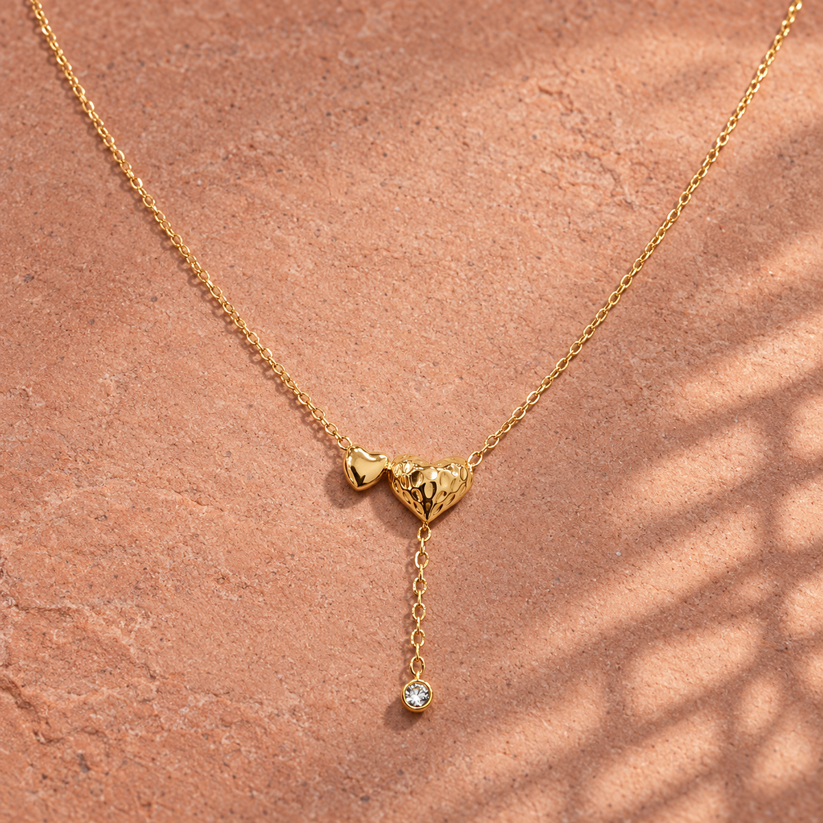 Gold-tone 316L stainless steel necklace with two heart pendants, one textured, and a drop chain ending in a clear crystal, lying on a stone surface