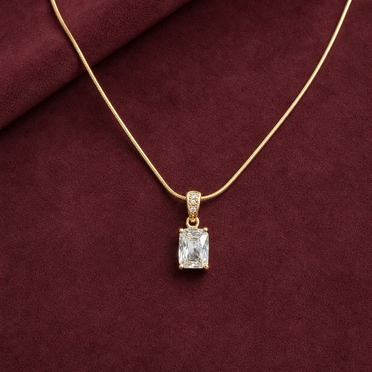 Gold-tone 316L stainless steel pendant necklace with a rectangular clear stone and pavé-set bail on a snake chain, displayed on maroon fabric