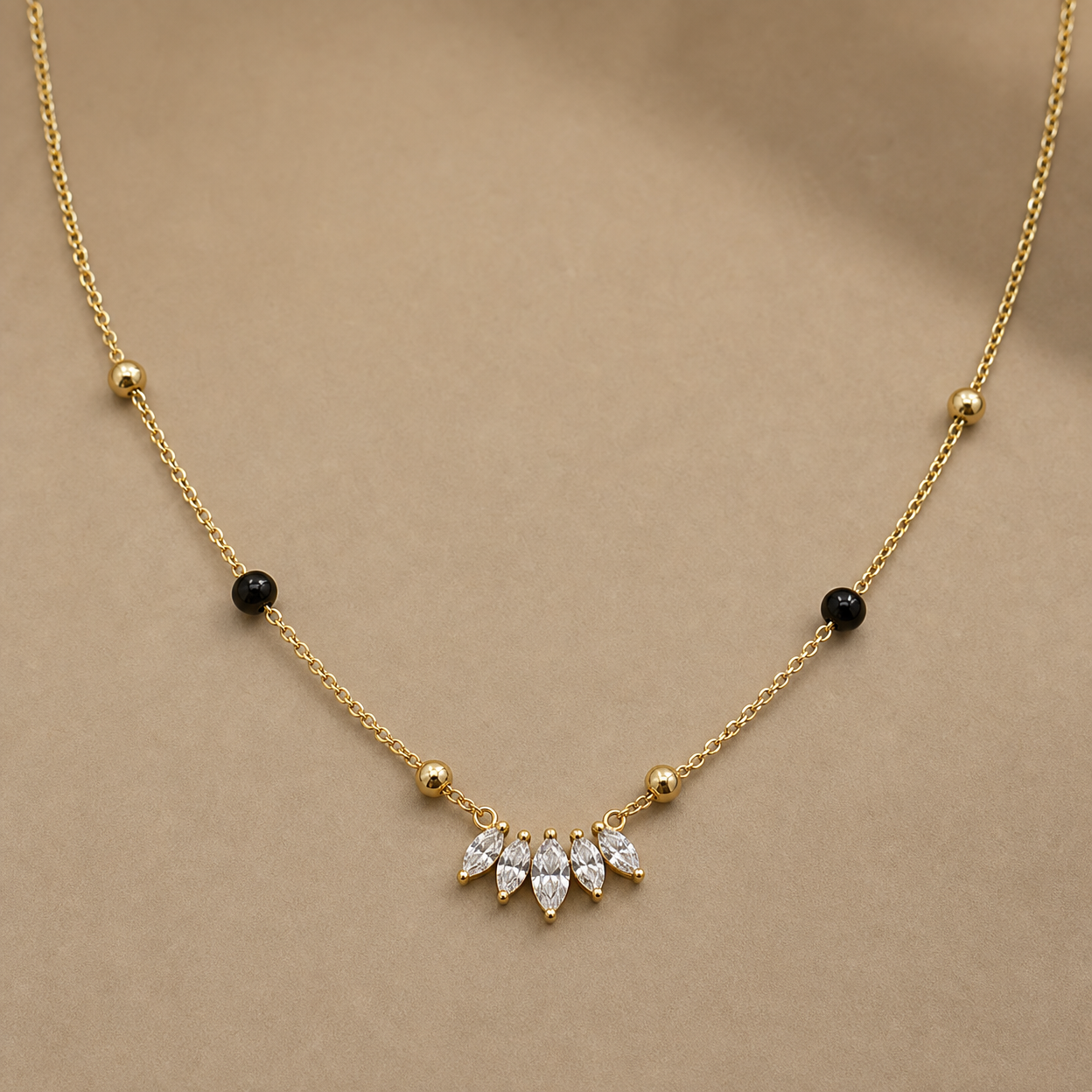 Gold-tone 316L steel necklace with a fine chain, black and gold beads, and a row of five marquise crystals at the centre, laid flat on a beige surface
