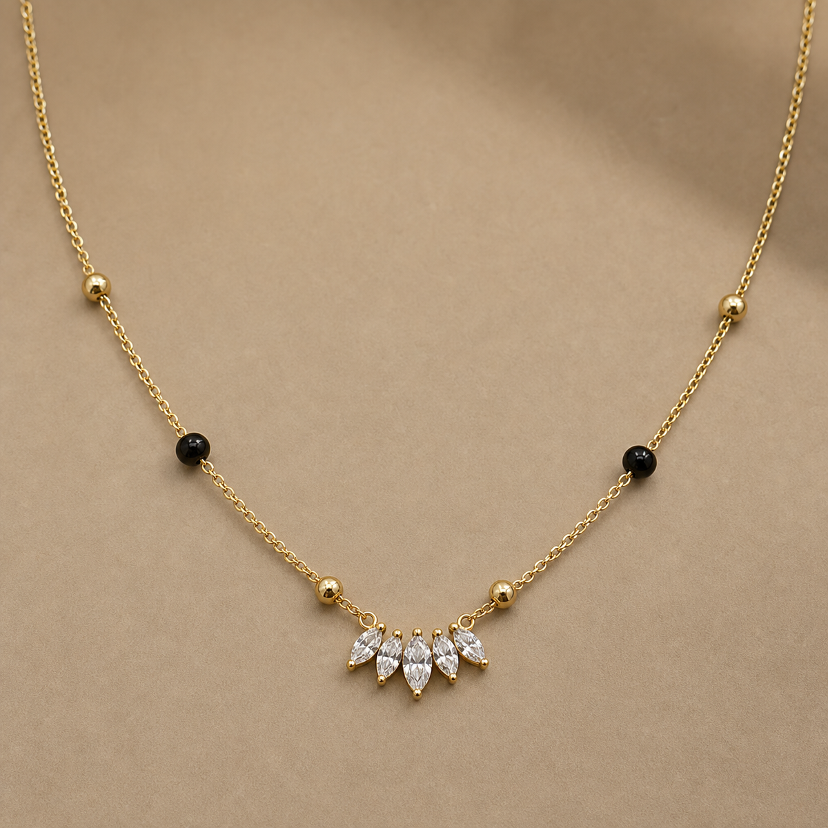Gold-tone 316L steel necklace with a fine chain, black and gold beads, and a row of five marquise crystals at the centre, laid flat on a beige surface