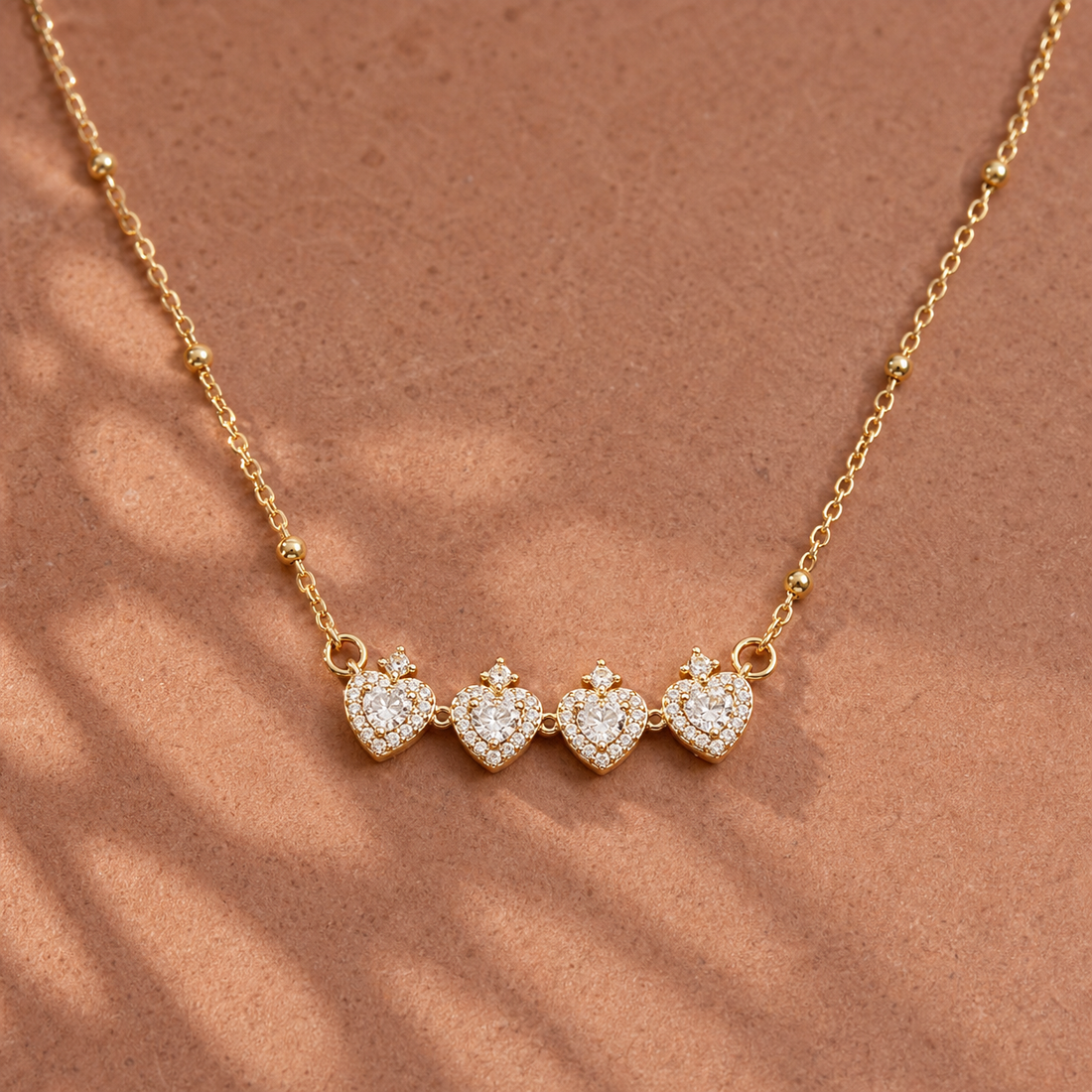 Gold-tone 304 stainless steel necklace with four heart pendants set with clear stones and a small crown stone on top, on a beaded chain, flat view