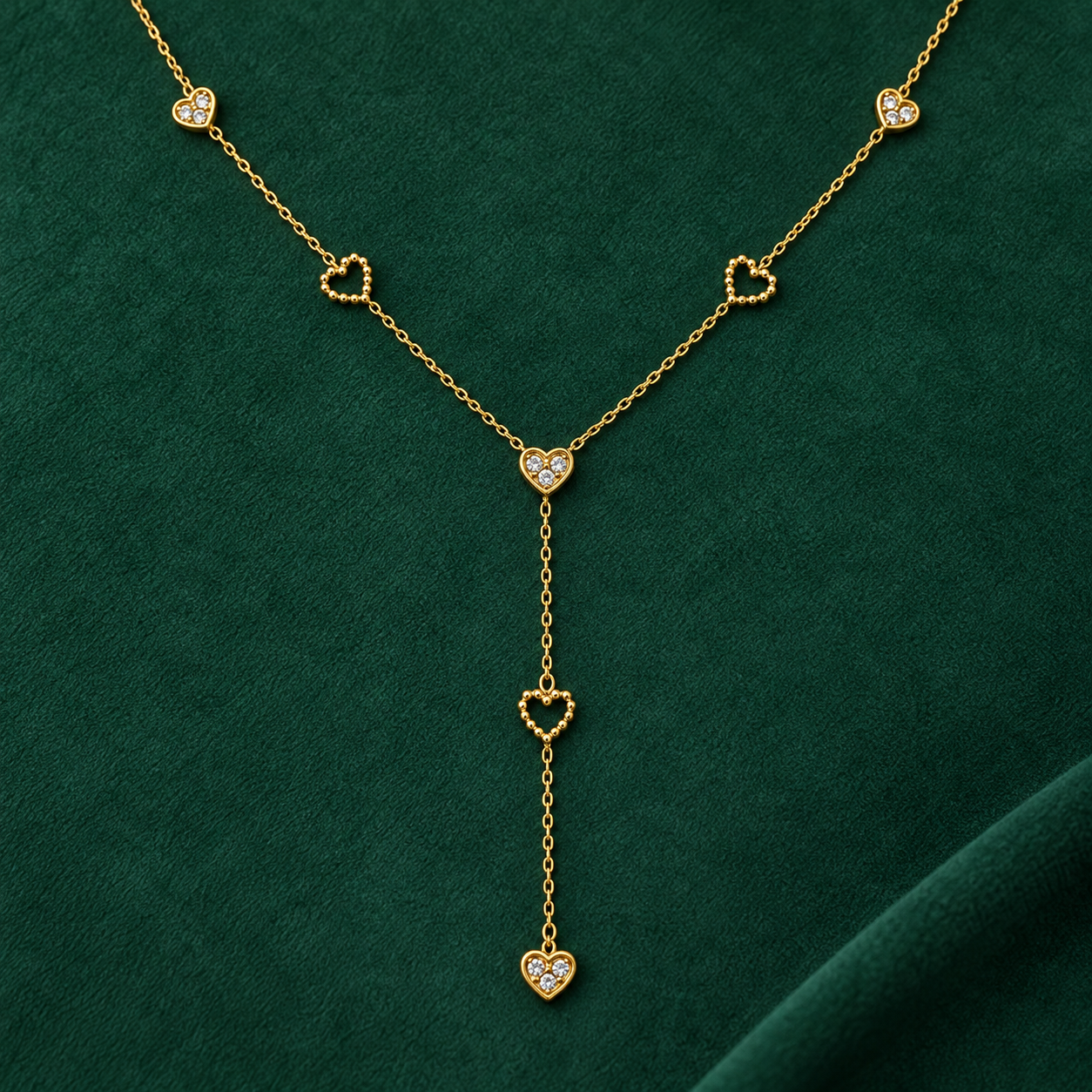 Gold-tone 316L stainless steel Y-drop necklace with heart motifs, some set with clear stones and some open beaded hearts, laid flat on green fabric