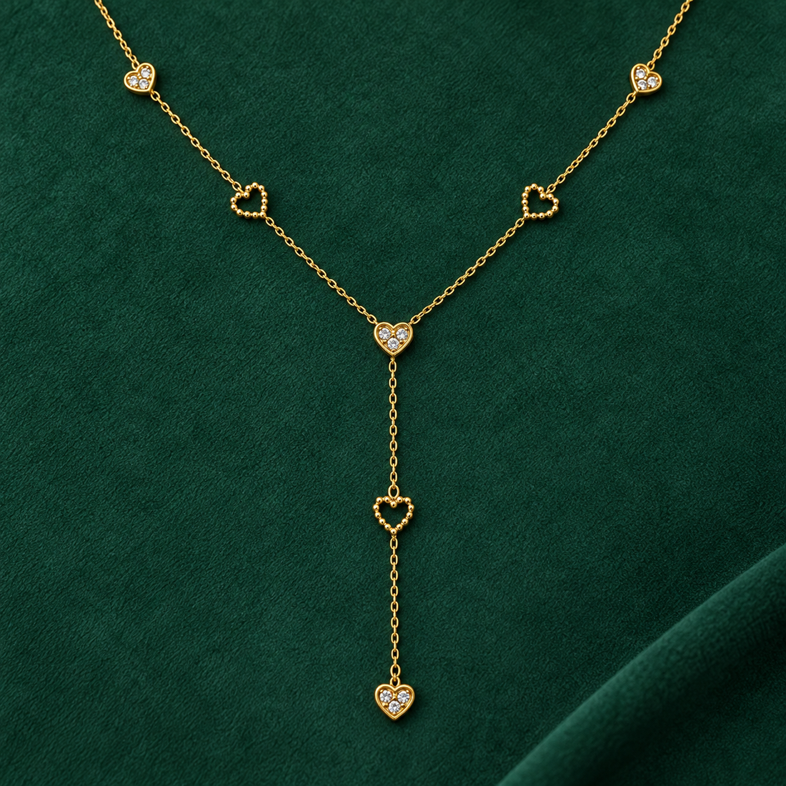 Gold-tone 316L stainless steel Y-drop necklace with heart motifs, some set with clear stones and some open beaded hearts, laid flat on green fabric