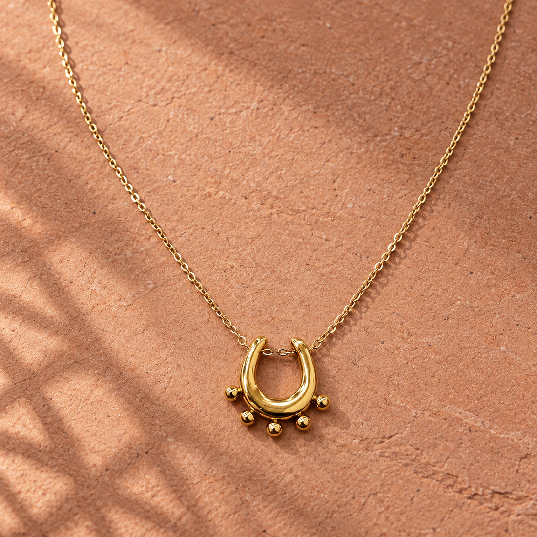 Gold-tone 316L stainless steel necklace with a thin cable chain and a U-shaped pendant edged with five small ball studs, laid on a sandy surface