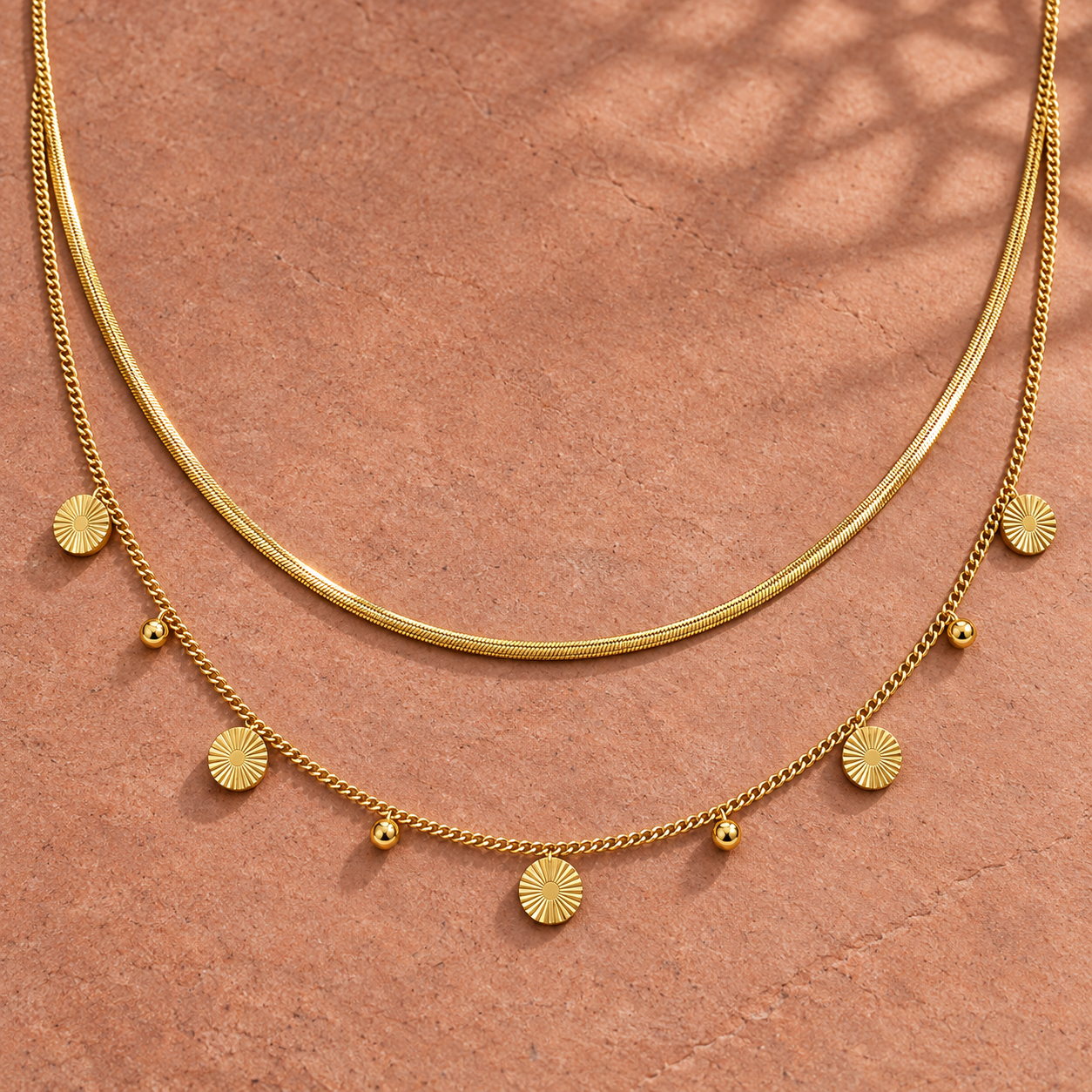 Gold-tone 316L stainless steel layered necklace set with a snake chain and a curb chain holding disc and ball charms, laid flat on a terracotta surface
