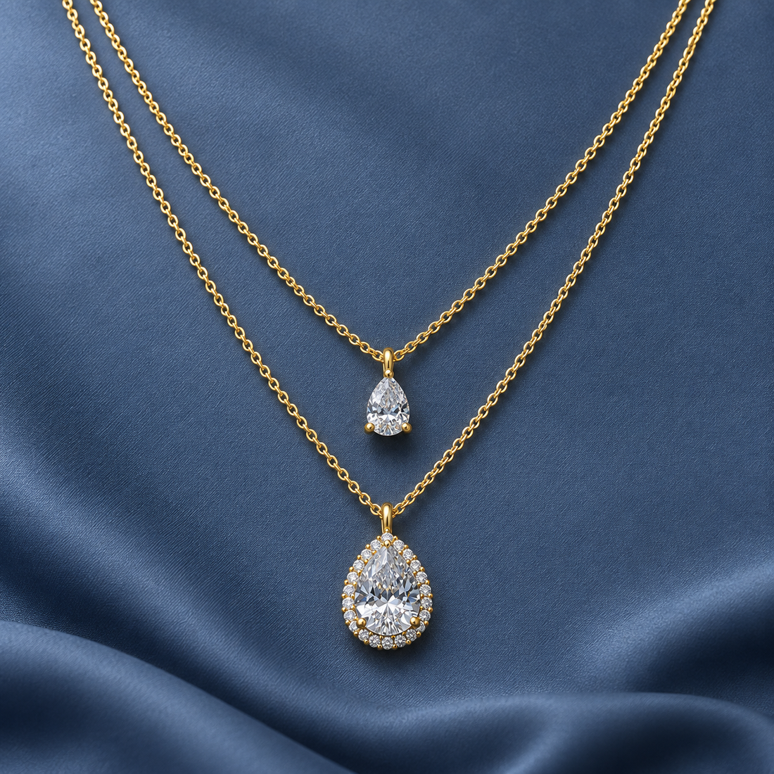 Gold-tone 316L stainless steel layered necklace with two pear-cut crystal pendants, one with a halo setting, on blue satin