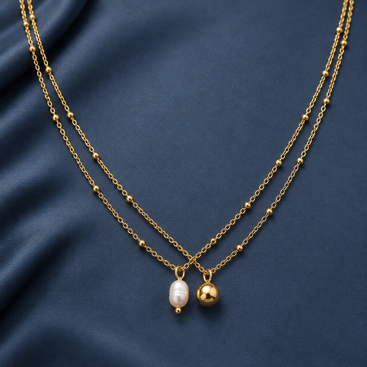 Gold-tone 304 stainless steel double-strand necklace with bead-accented chains and a white pearl drop and a plain gold-tone ball charm, on a dark blue fabric