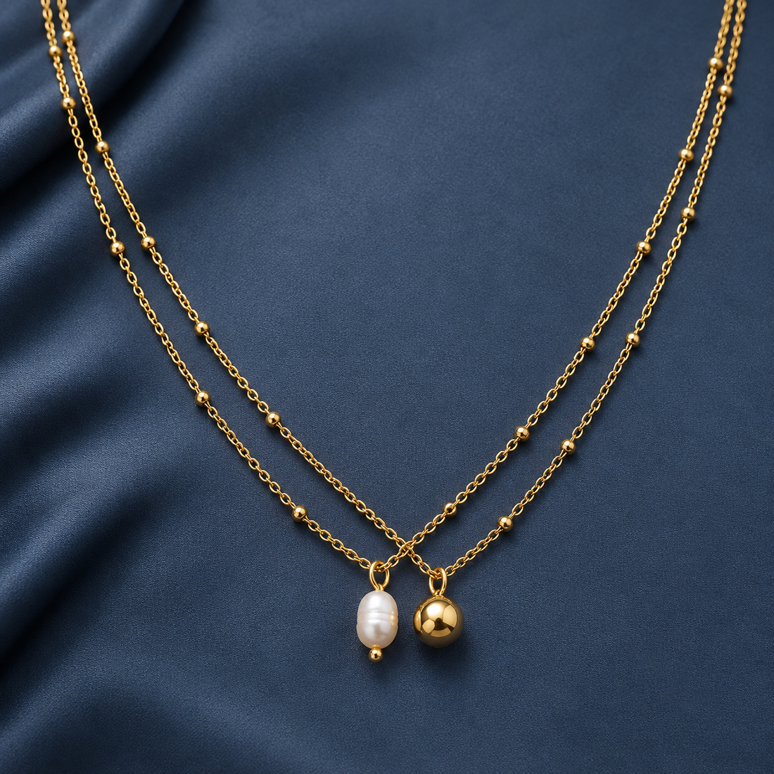 Gold-tone 304 stainless steel double-strand necklace with bead-accented chains and a white pearl drop and a plain gold-tone ball charm, on a dark blue fabric