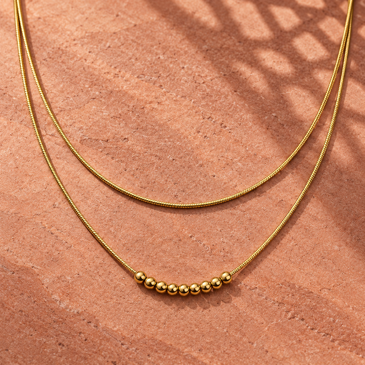 Gold-tone 304 stainless steel double-strand necklace with a fine snake chain and a row of round beads at the front, laid flat on a terracotta surface