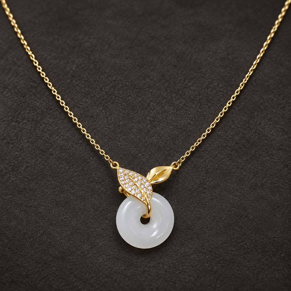 Gold-tone 316L stainless steel necklace with a white jade-look donut pendant and a leaf-shaped crystal-set bail on a fine cable chain, laid flat on dark fabric
