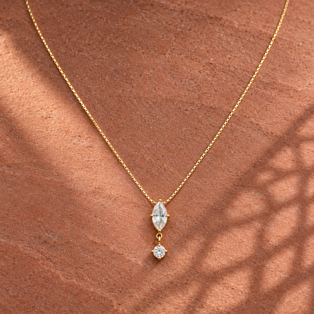 Gold-tone 304 stainless steel necklace with a thin box chain and a marquise and round clear stone drop pendant, laid on a stone surface