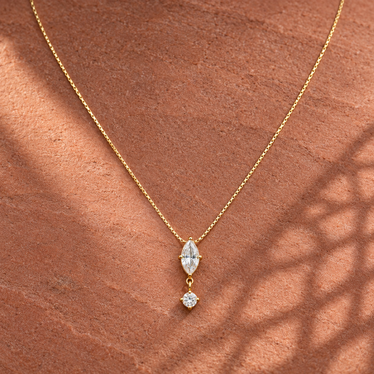 Gold-tone 304 stainless steel necklace with a thin box chain and a marquise and round clear stone drop pendant, laid on a stone surface