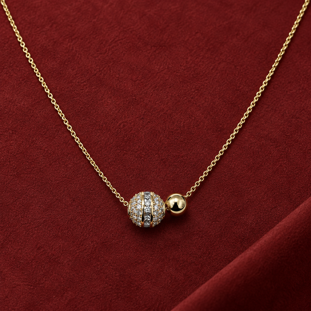 Gold-tone 316L stainless steel necklace with a fine cable chain and a pavé-set ball pendant beside a plain gold-tone bead, shown on a red surface