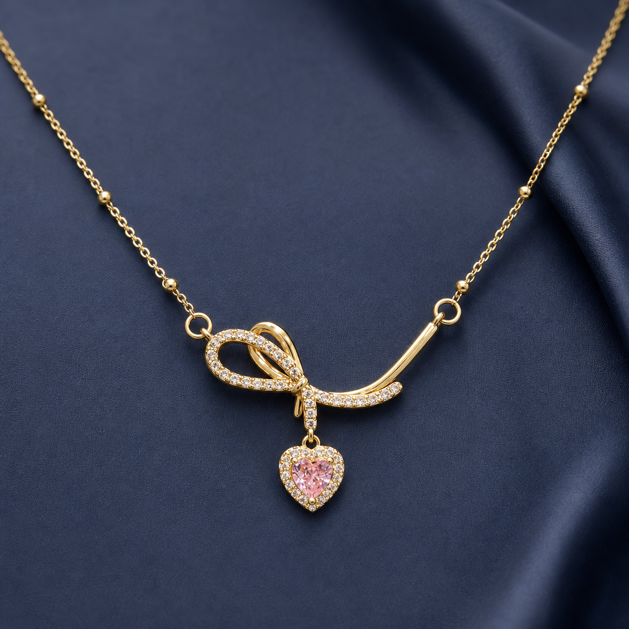 Gold-tone 304 stainless steel necklace with a bow pendant set with clear stones and a pink heart-shaped drop, on a dark blue fabric backdrop