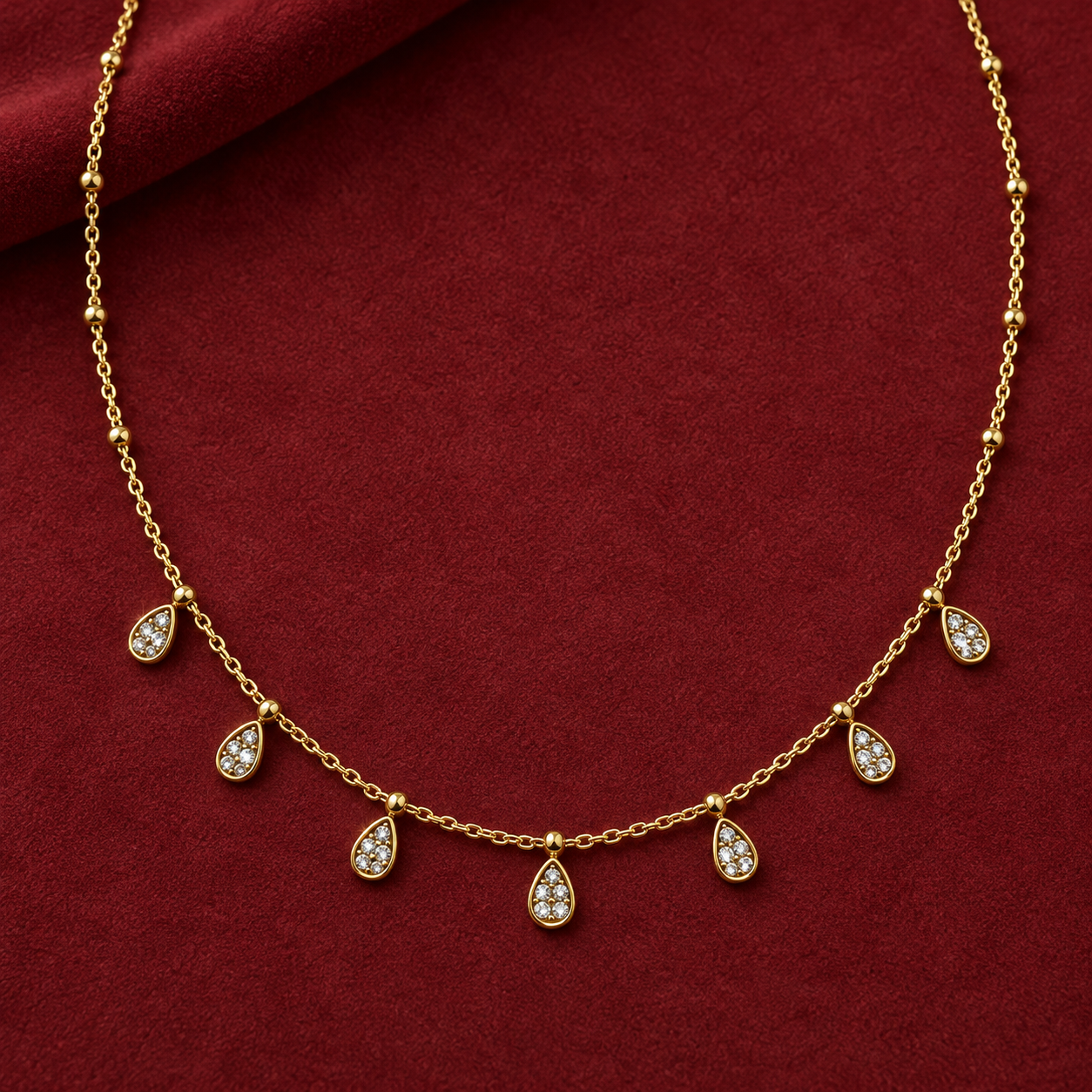 Gold-tone 316L stainless steel necklace with a fine cable chain, bead accents and seven teardrop pendants set with clear stones, laid on a red cloth