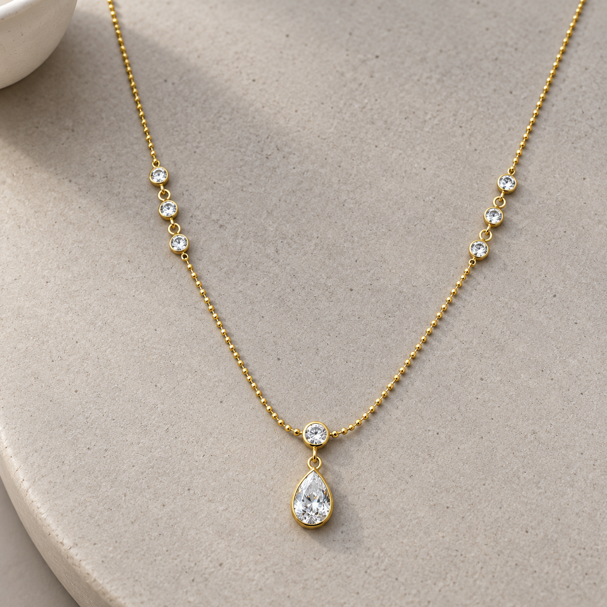 Gold-tone 316L stainless steel ball-chain necklace with round clear stones and a pear-cut clear stone pendant, lying flat on a grey surface