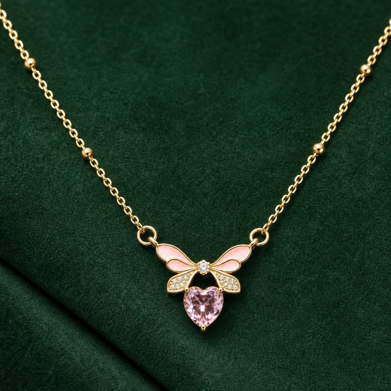 Gold-tone 304 stainless steel necklace with a pink enamel bow and a pink heart-cut stone, shown lying on green fabric