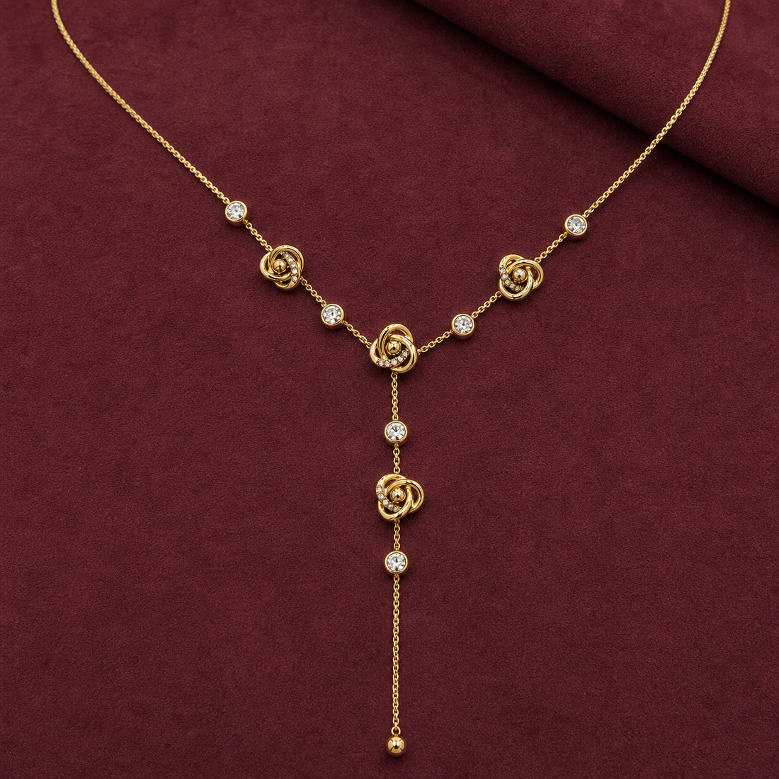 Gold-tone 316L stainless steel Y-drop necklace with knot-rose motifs and clear stone-set links on a fine chain, displayed flat on maroon fabric