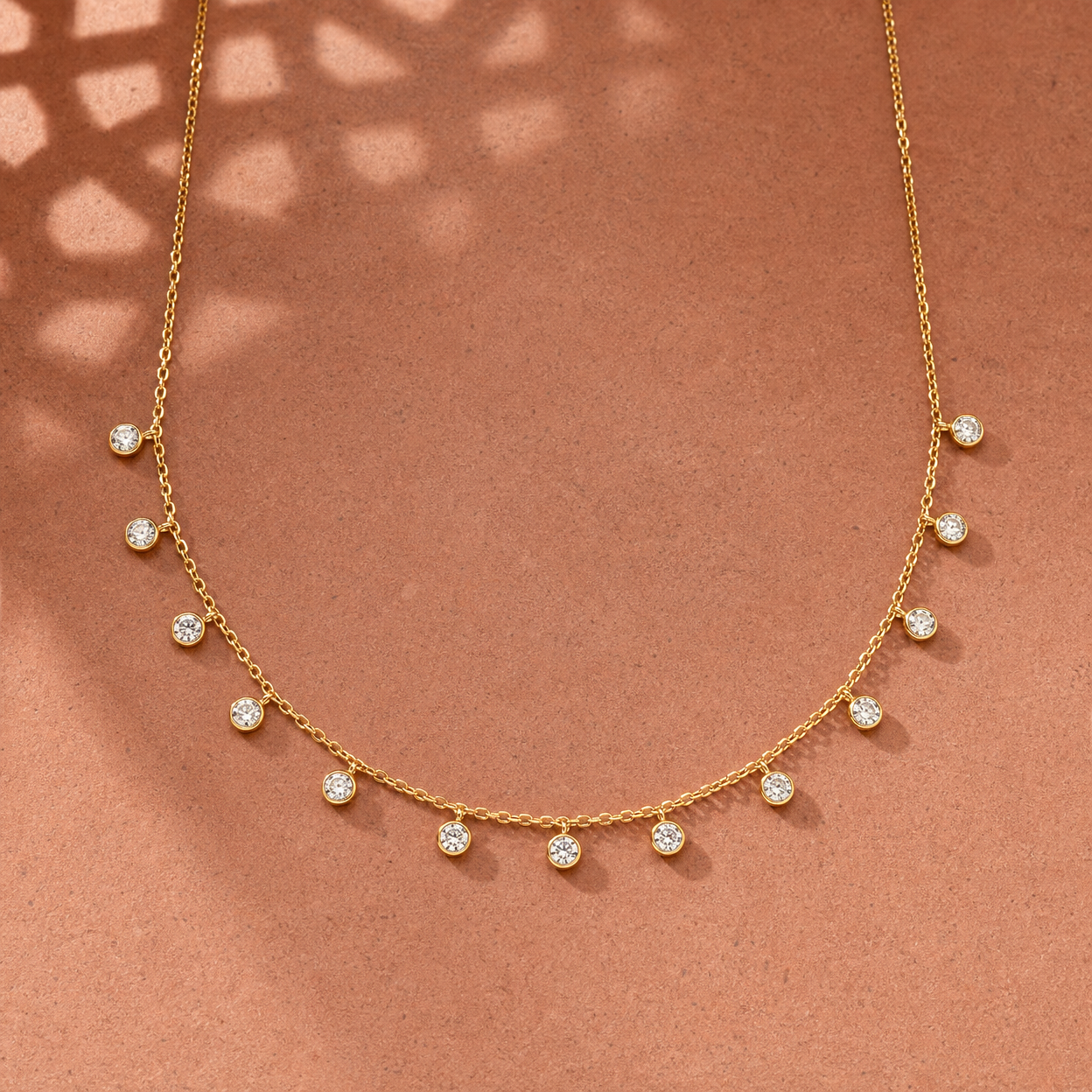 Gold-tone 304 stainless steel necklace with a fine cable chain and thirteen bezel-set clear stones dangling from it, laid flat on a terracotta surface