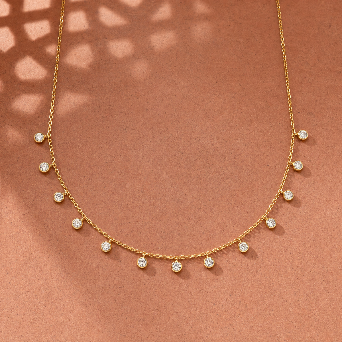 Gold-tone 304 stainless steel necklace with a fine cable chain and thirteen bezel-set clear stones dangling from it, laid flat on a terracotta surface