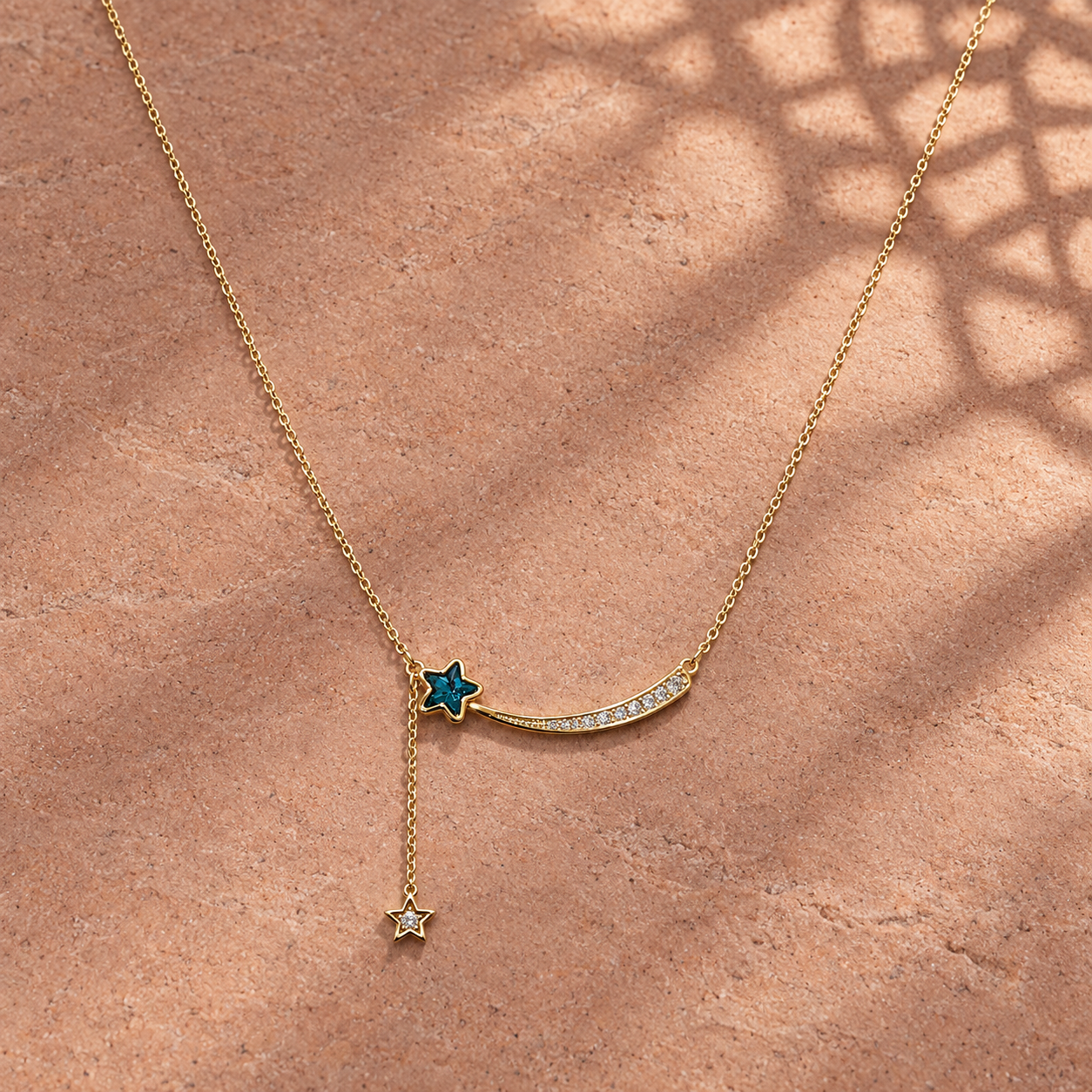 Gold-tone 316L stainless steel necklace with a curved shooting-star pendant set with clear stones and a blue star charm on a side drop, lying on stone