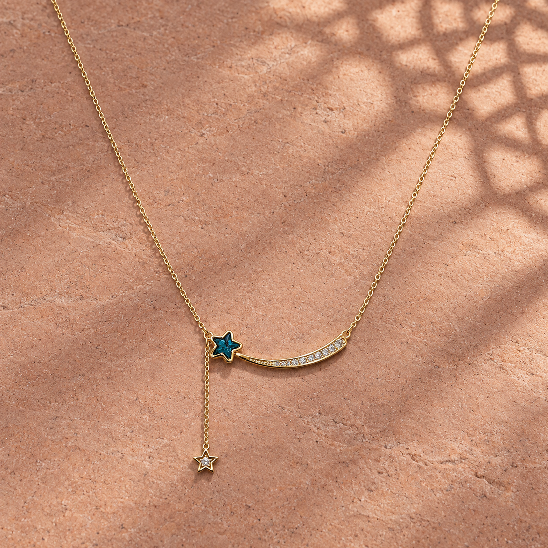 Gold-tone 316L stainless steel necklace with a curved shooting-star pendant set with clear stones and a blue star charm on a side drop, lying on stone