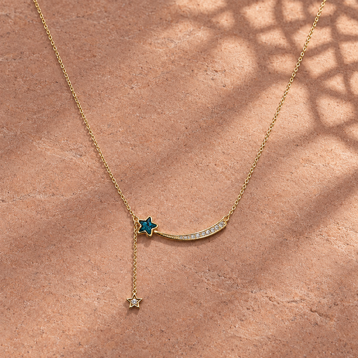 Gold-tone 316L stainless steel necklace with a curved shooting-star pendant set with clear stones and a blue star charm on a side drop, lying on stone