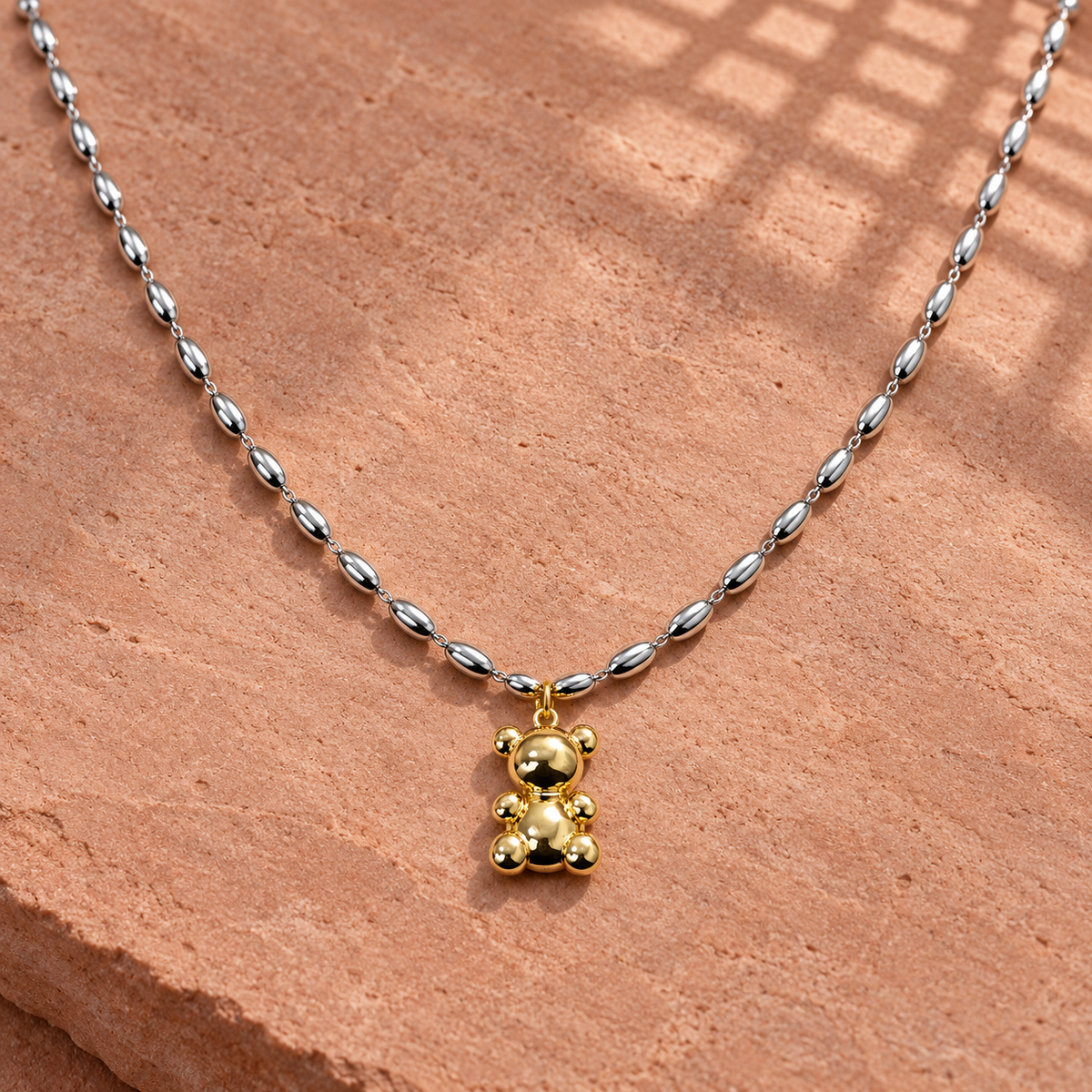 Two-tone 316L stainless steel necklace with an oval bead chain and a gold-tone teddy bear pendant with ball-shaped limbs, laid flat on a stone surface