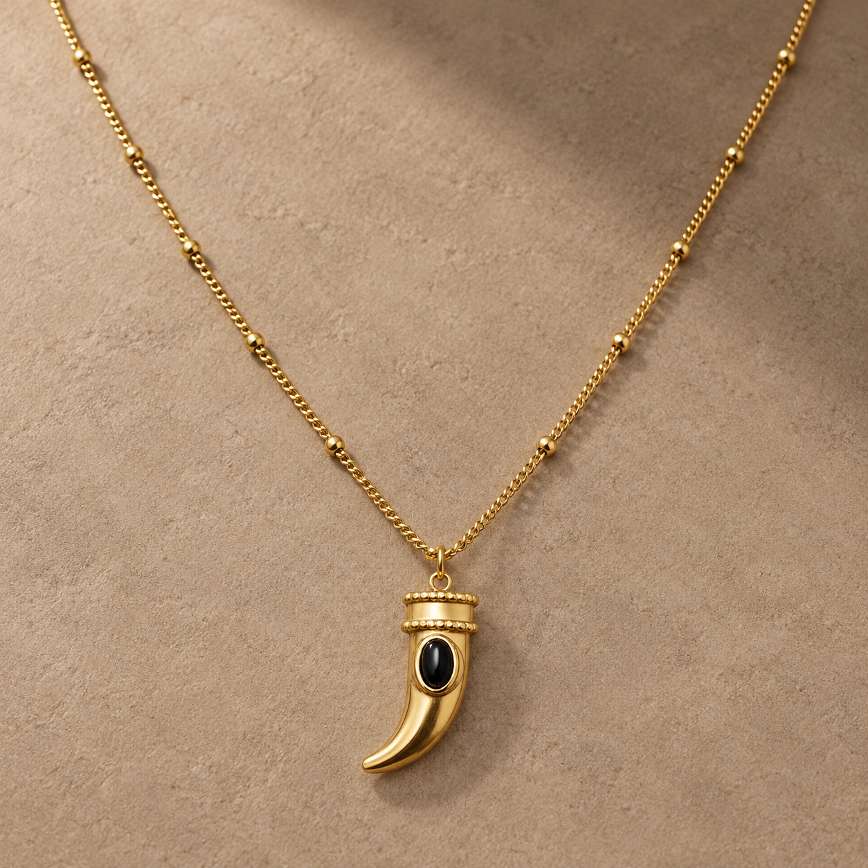 Gold-tone 304 stainless steel necklace with a curved horn pendant set with a black oval stone on a beaded chain, lying flat on a stone surface
