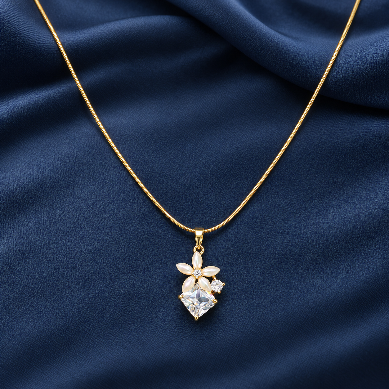 Gold-tone 316L pendant necklace with a snake chain and a flower pendant of pearl-like petals around a square clear stone, laid on dark blue satin