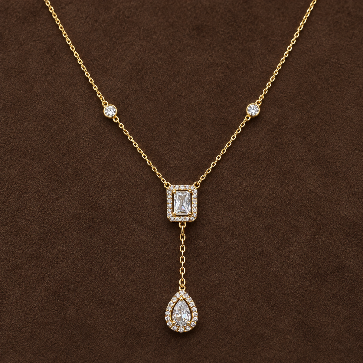 Gold-tone 316L stainless steel necklace with a clear stone-set rectangle and teardrop pendant on a Y-drop chain, laid flat on a brown surface