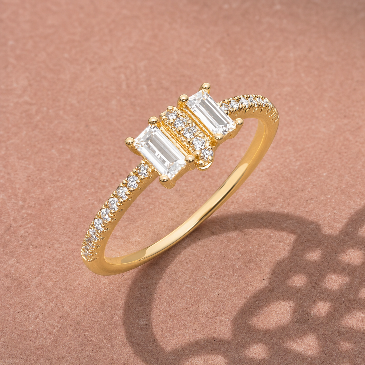 Gold-tone 316L ring with three baguette-cut white stones and a row of small round stones along the band, on a rose-coloured surface