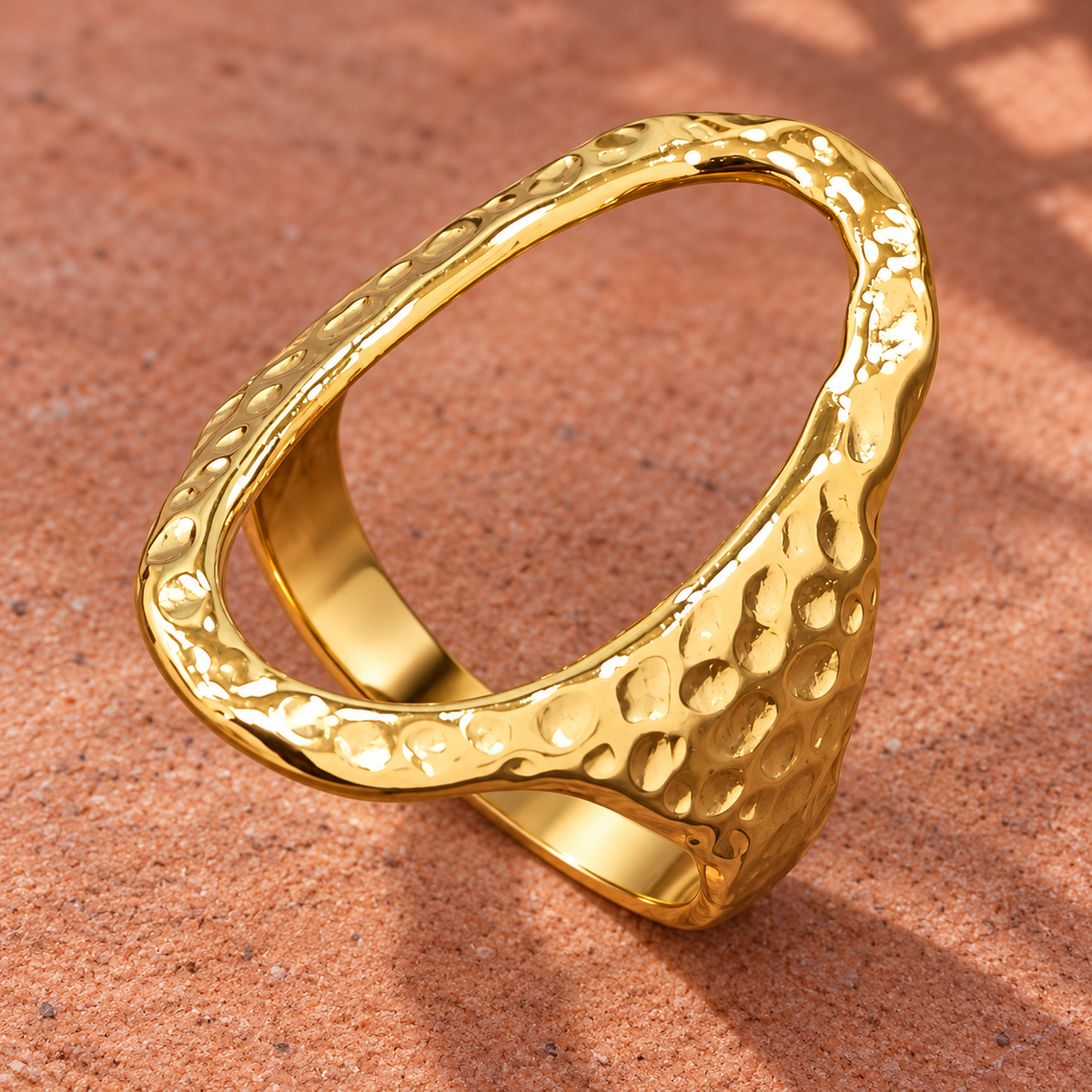 Gold-tone 304 stainless steel open oval ring with a hammered texture on a wide band, shown on a sandy surface