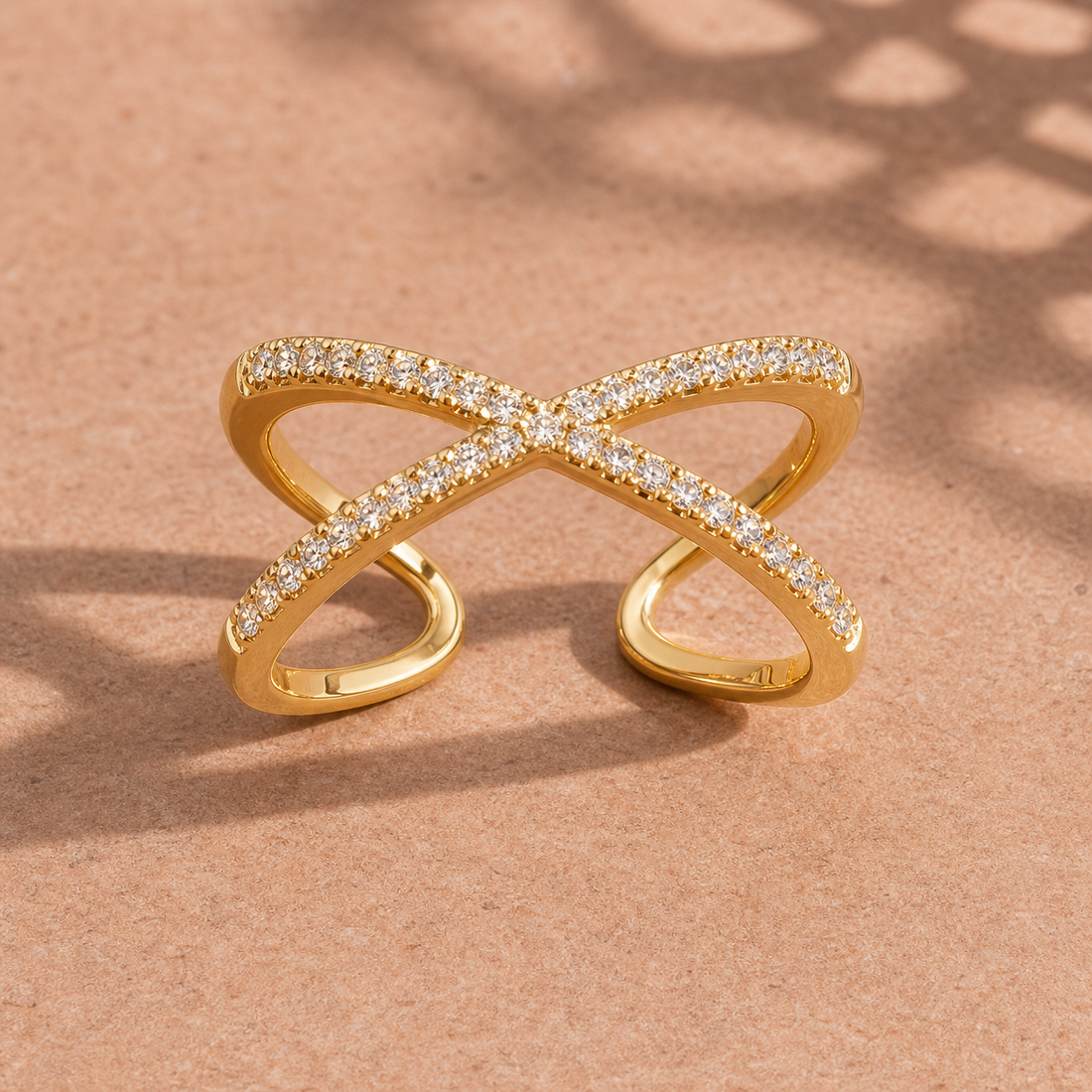 Gold-tone 304 stainless steel crossover ring with a criss-cross band set with clear stones, shown lying on a sandy surface