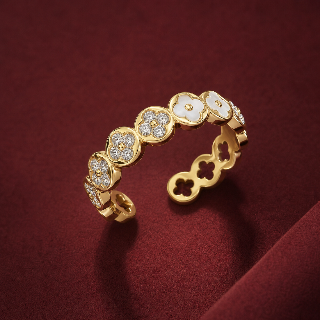 Gold-tone 316L stainless steel open band ring with clover motifs alternating white enamel and crystal pavé, shown on a dark red surface