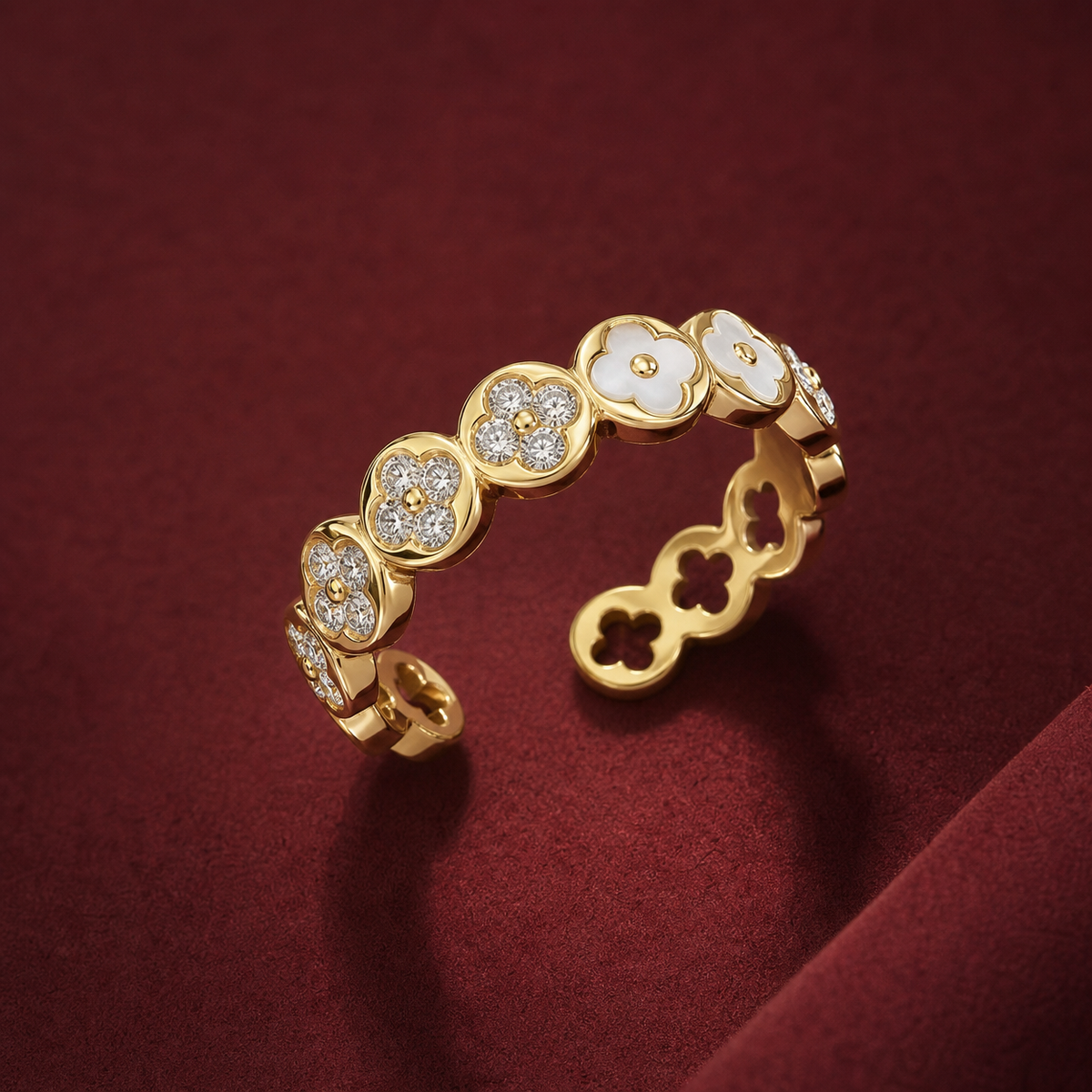 Gold-tone 316L stainless steel open band ring with clover motifs alternating white enamel and crystal pavé, shown on a dark red surface