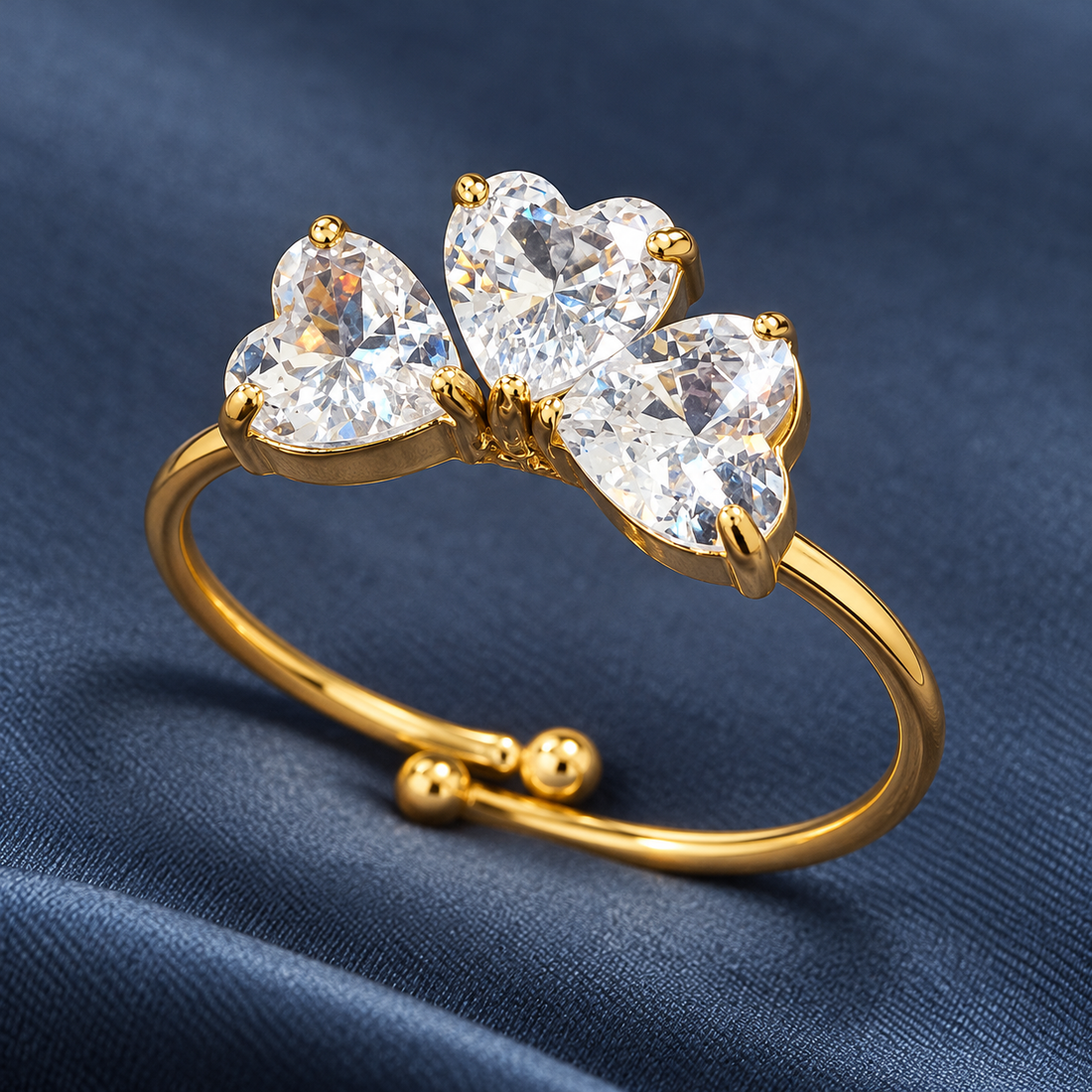 Gold-tone 304 stainless steel open ring with three heart-cut clear crystals in prong settings and ball-end band ends, shown on a blue fabric
