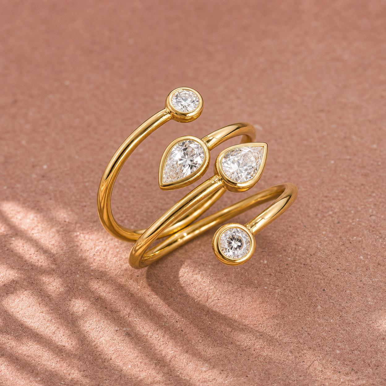 Gold-tone 316L stainless steel wrap ring with three round and two pear-cut clear stones in bezel settings, shown on a pink surface