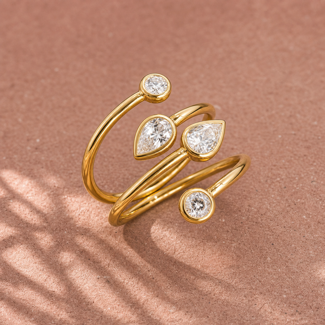 Gold-tone 316L stainless steel wrap ring with three round and two pear-cut clear stones in bezel settings, shown on a pink surface